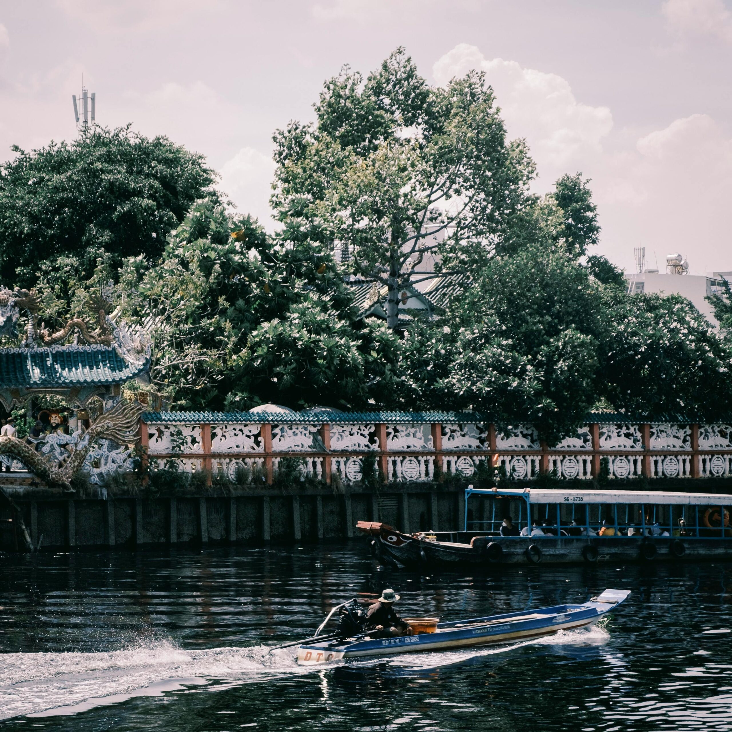 A speedboat cruises along a river, framed by lush greenery and an ornate temple wall, under a partly cloudy sky.