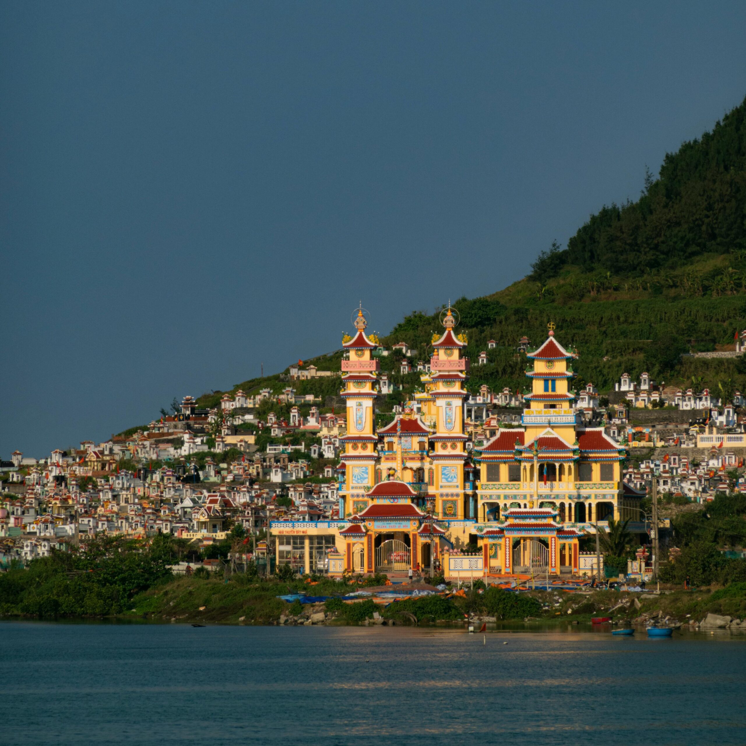 Vibrant Cao Dai temple on a riverside with hillside cemetery in Vietnam.