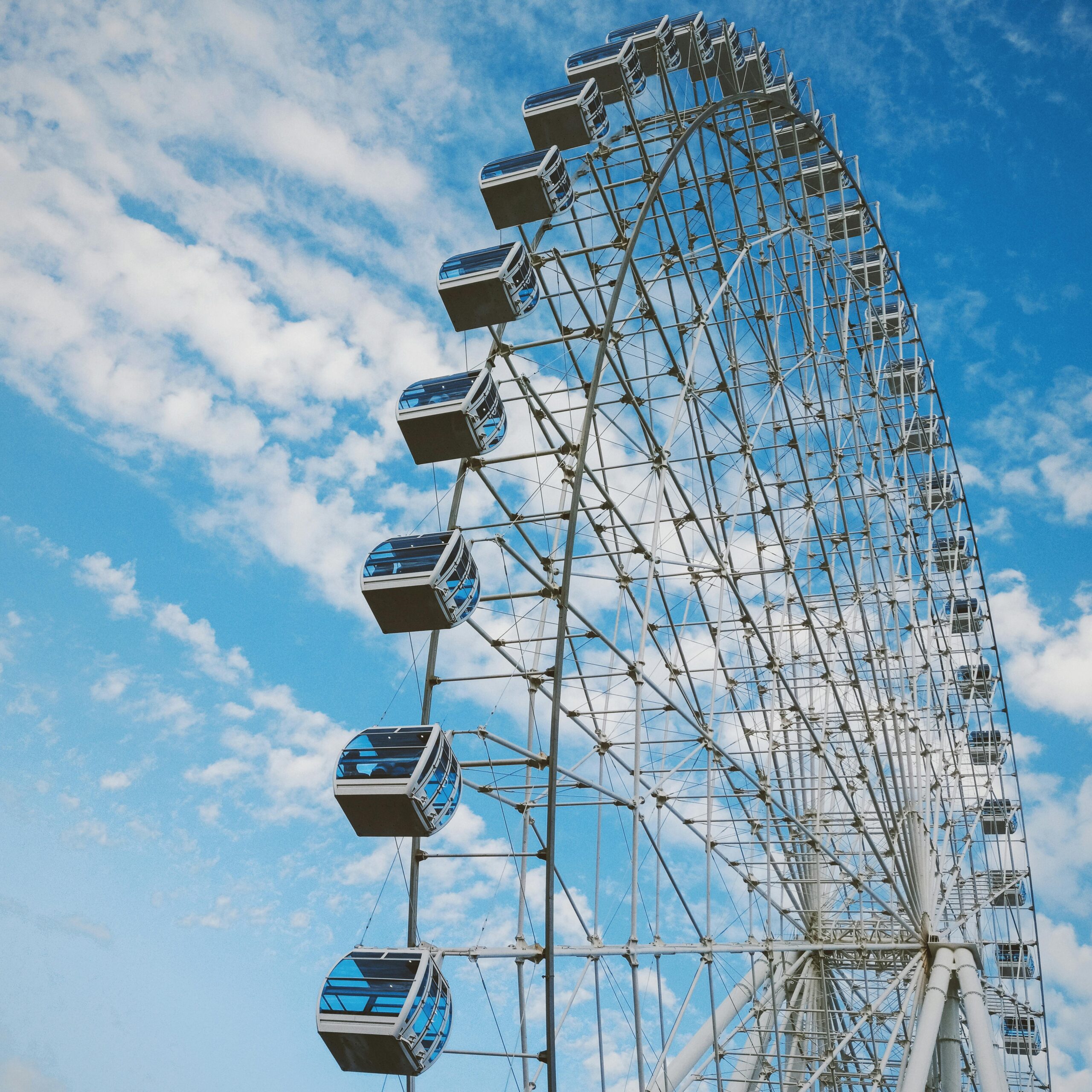 From below of big wheel with metal spokes and cabins under cloudy blue sky in fun park