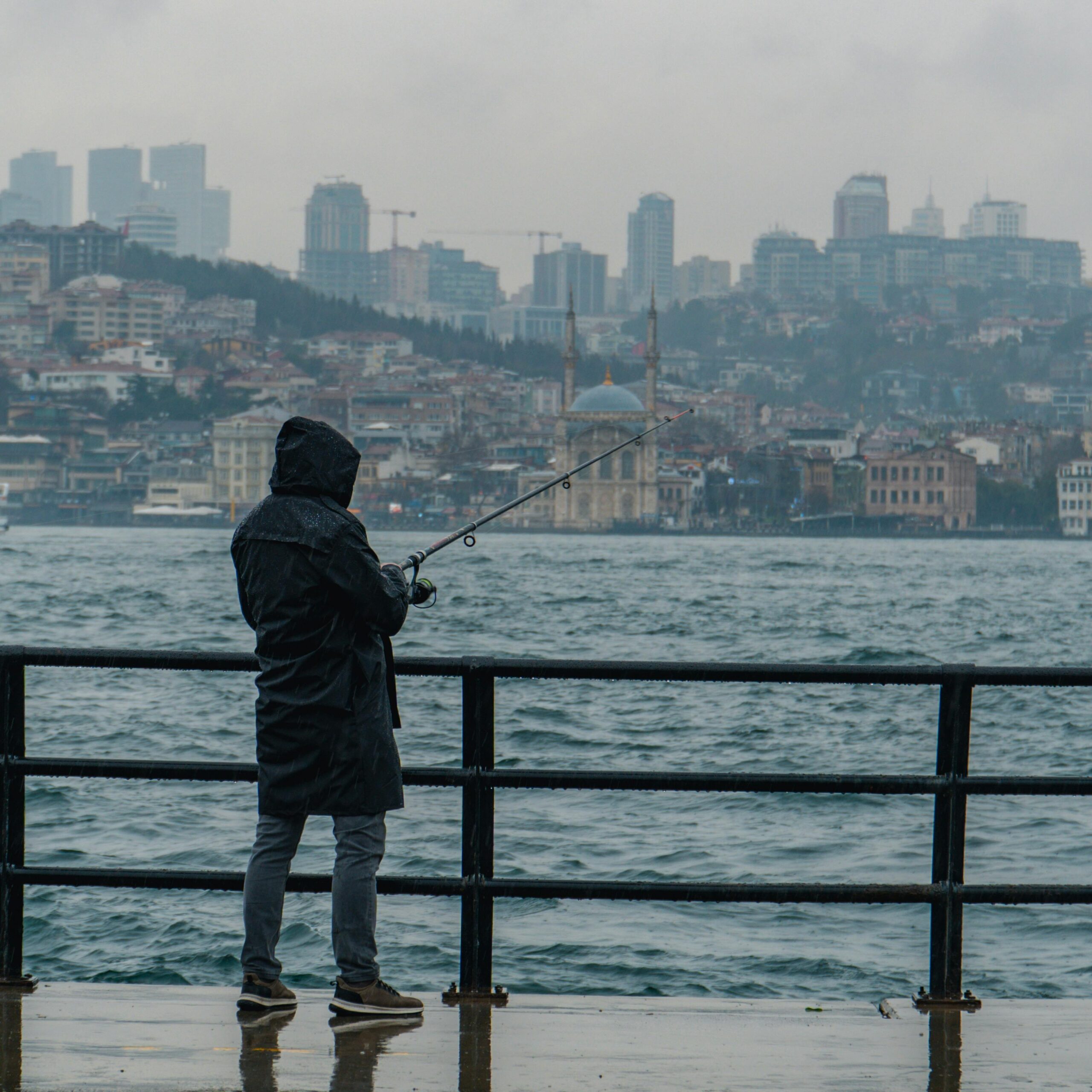 A lone fisherman casts a line by the city waterfront during a rainy day.