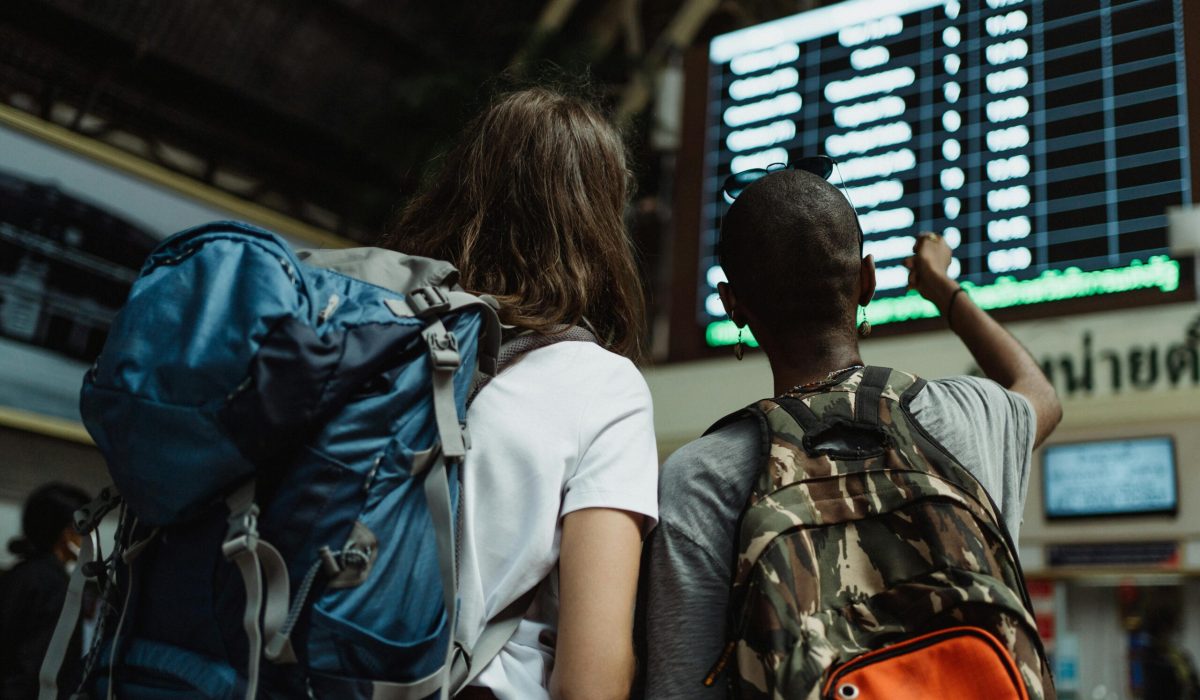 Two backpackers checking the digital board at a train station for travel information.
